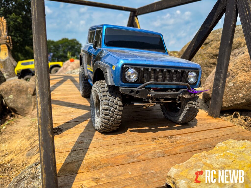 Blue RC off-road vehicle crossing a wooden bridge outdoors with rocks and a yellow vehicle in the background.