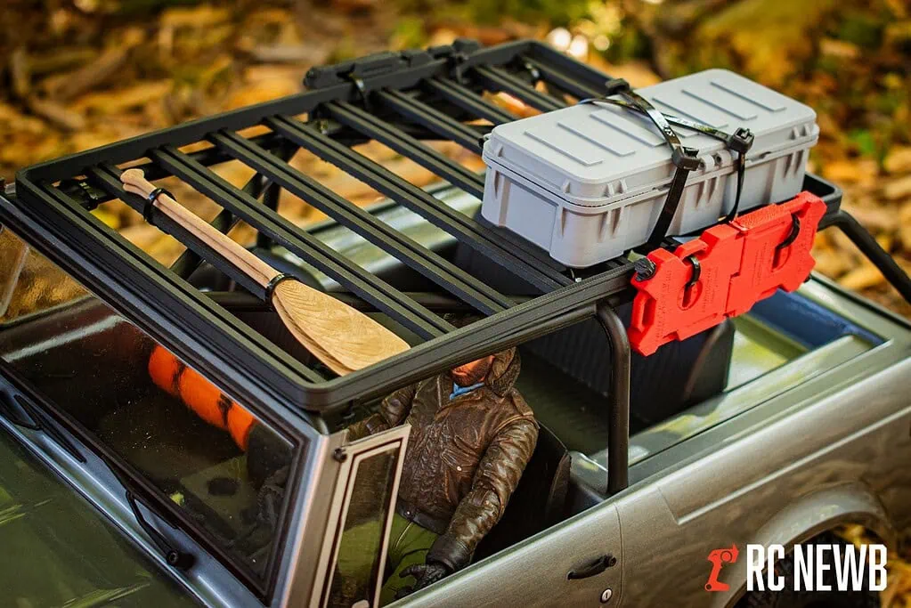 An RC car roof rack with outdoor gear including a paddle, storage box, and safety items, set against autumn leaves, demonstrating off-road RC vehicle customization and outdoor adventure preparedness.
