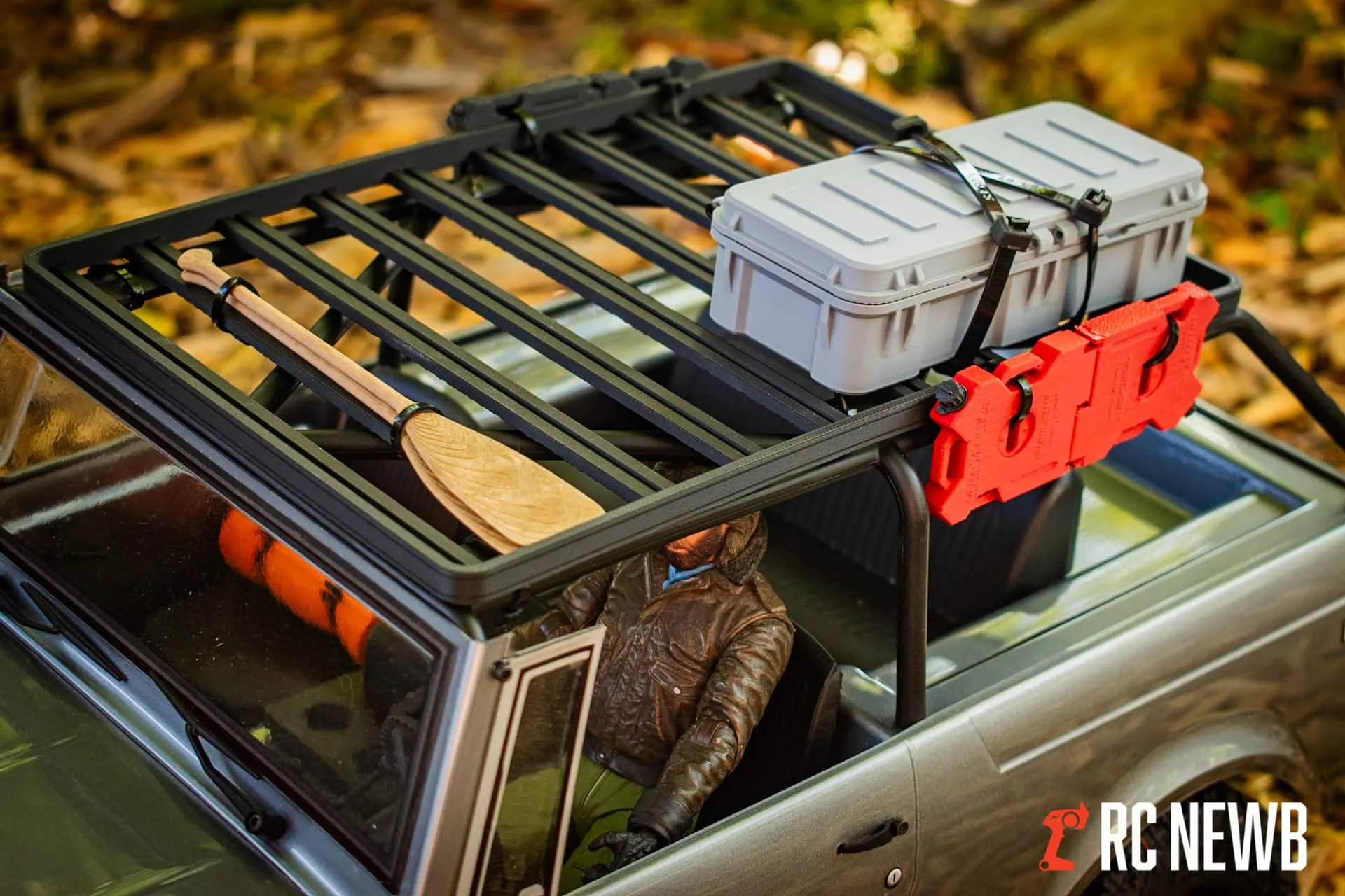 An RC car roof rack with outdoor gear including a paddle, storage box, and safety items, set against autumn leaves, demonstrating off-road RC vehicle customization and outdoor adventure preparedness.