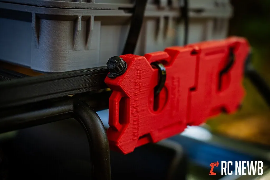 Red 3D-printed RC car part mounted on a black frame with a gray storage box in the background, showcasing DIY RC hobby modifications.