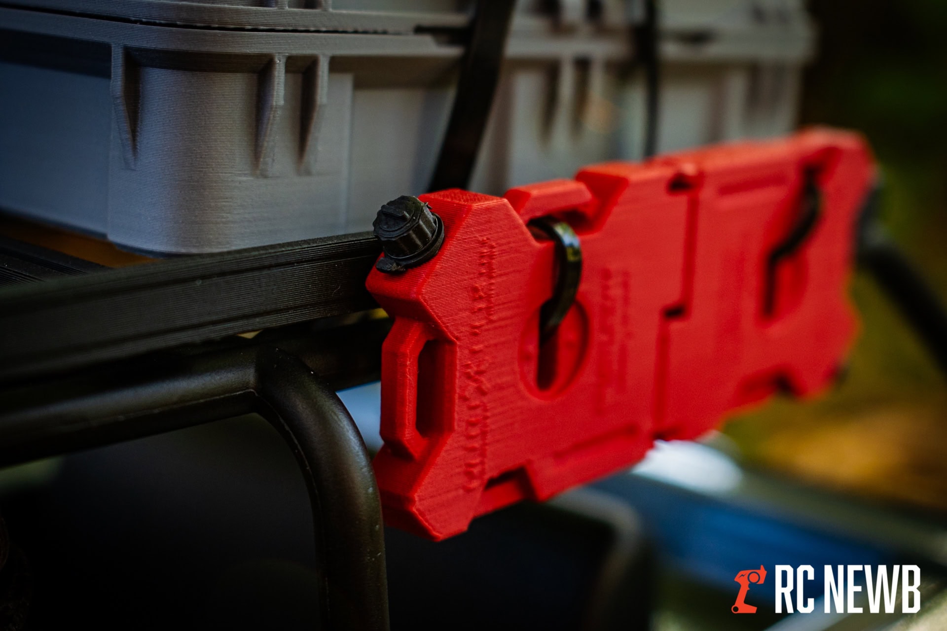 Red 3D-printed RC car part mounted on a black frame with a gray storage box in the background, showcasing DIY RC hobby modifications.