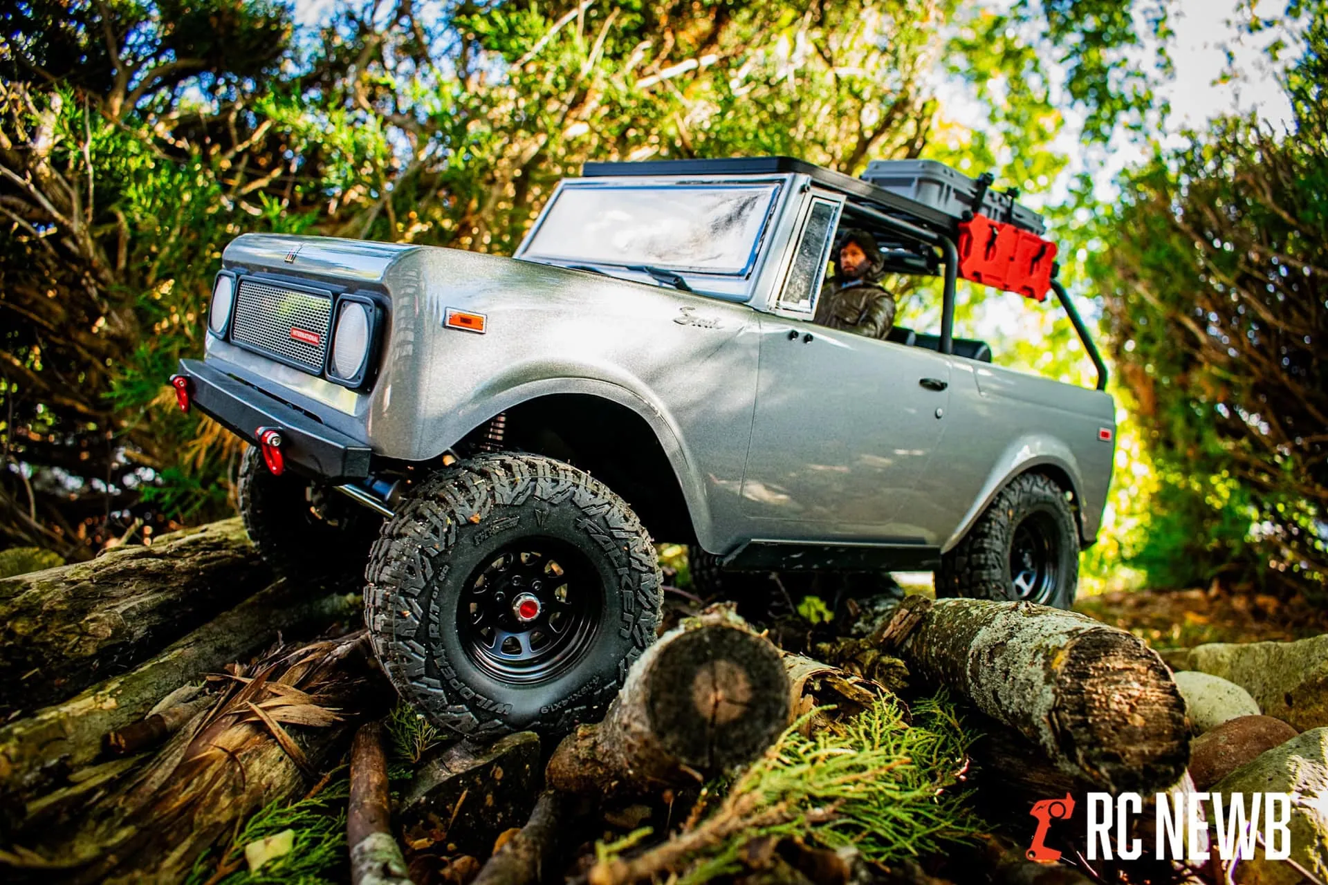 Off-road RC truck navigating over logs in a forest setting during daytime.